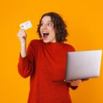 europejska kobieta nosi sweter za pomocą srebrnego laptopa i karty kredytowej, stojąc na białym tle na żółto