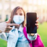 Młoda pewna studentka kobieta w masce medycznej wskazuje na pusty ekran smartfona i stoi na kampusie uniwersyteckim