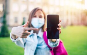 Młoda pewna studentka kobieta w masce medycznej wskazuje na pusty ekran smartfona i stoi na kampusie uniwersyteckim