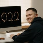 Praca zdalna. Biznesmen pracuje zdalnie, korzystając z komputera typu „wszystko w jednym”. Uśmiechnięty facet pracuje z domu w pobliżu wyświetlacza z 2022 na nim.