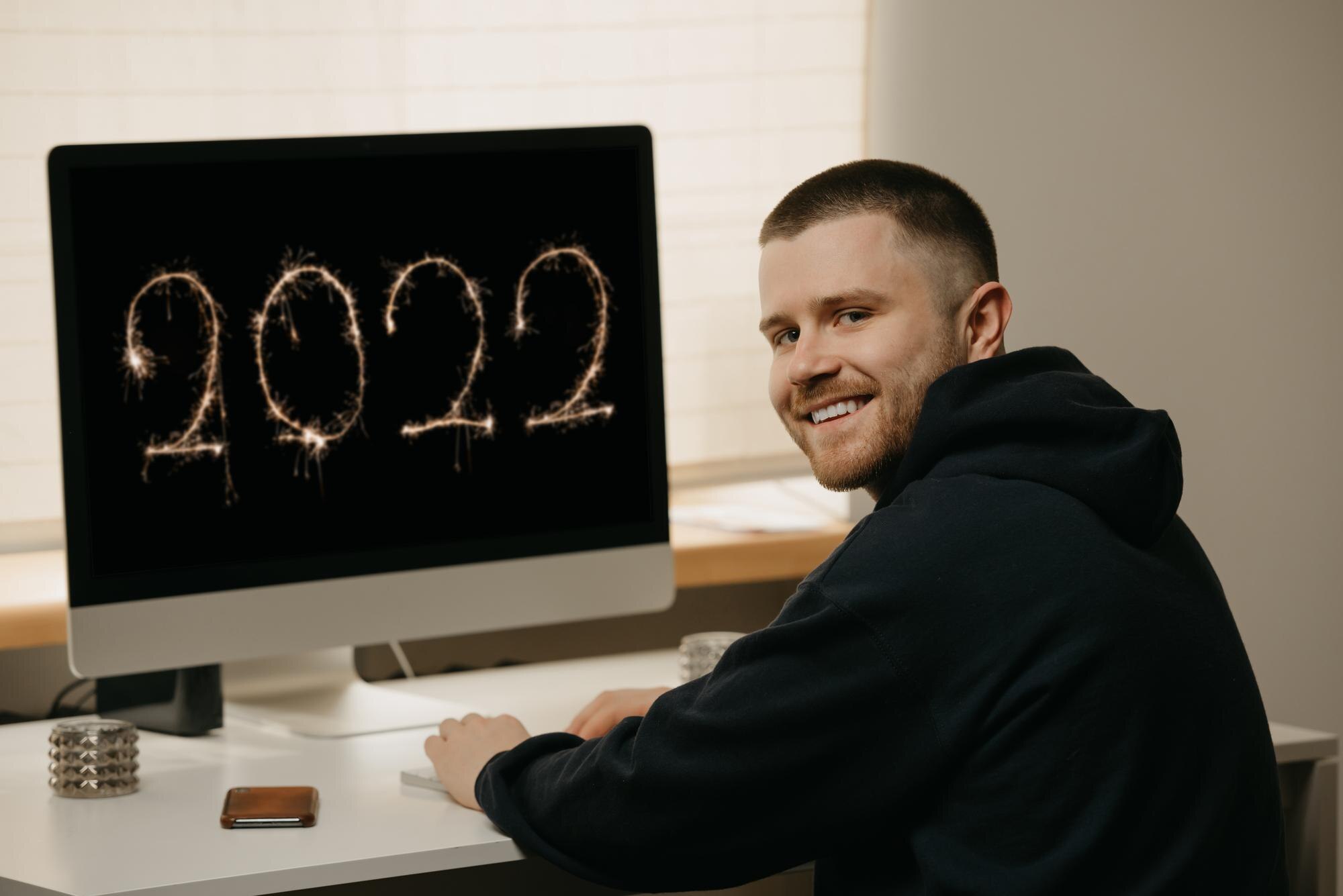 Praca zdalna. Biznesmen pracuje zdalnie, korzystając z komputera typu „wszystko w jednym”. Uśmiechnięty facet pracuje z domu w pobliżu wyświetlacza z 2022 na nim.