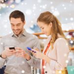 sprzedaż, konsumpcjonizm, technologia i koncepcja ludzi - szczęśliwa para z torbami na zakupy i smartfonami wysyłającymi SMS-y w centrum handlowym z efektem śniegu