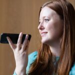 W biurze kobieta z smartfonem uśmiecha się i mówi do urządzenia, angażując się w rozmowę