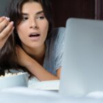 Młoda kobieta używa laptop i jedzący popkorn